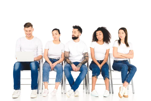 Homem alegre usando laptop enquanto cansado grupo multiétnico de pessoas sentadas em cadeiras isoladas em branco — Fotografia de Stock