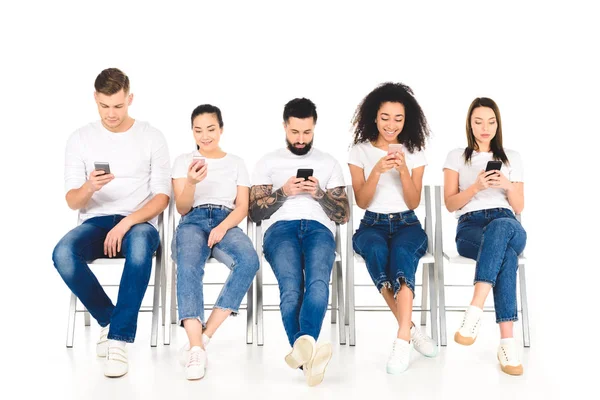 Multicultural group of young people sitting on chairs with smartphones isolated on white — Stock Photo