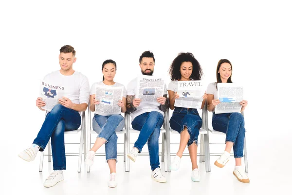 Grupo multiétnico de jovens sentados em cadeiras com pernas cruzadas e lendo jornais isolados em branco — Fotografia de Stock