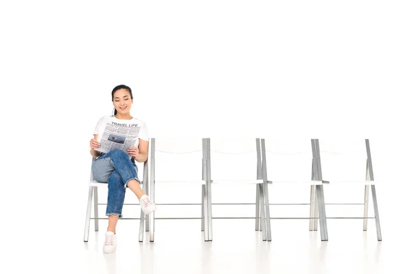 Sorrindo menina sentada na cadeira com pernas cruzadas e lendo jornal isolado no branco — Fotografia de Stock