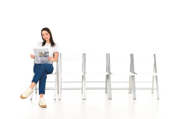 Attractive girl sitting on chair with crossed legs and reading newspaper isolated on white — Stock Photo