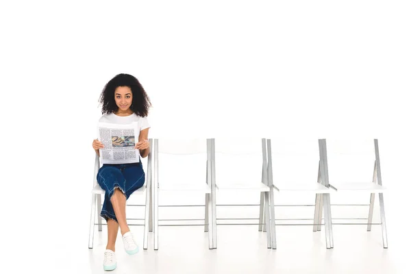 African american girl sitting on chair with crossed legs and newspaper while looking at camera isolated on white — Stock Photo