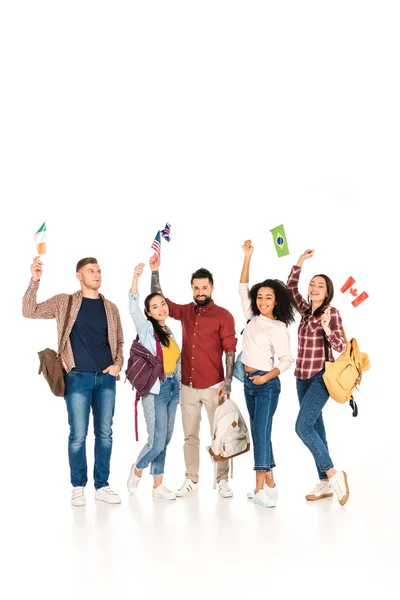 Glückliche multiethnische Gruppe von Menschen, die mit Rucksäcken und Flaggen verschiedener Länder über Köpfen isoliert auf weiß stehen — Stockfoto