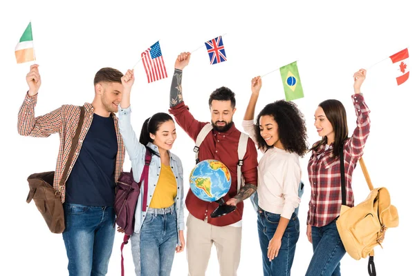 Gruppo multietnico di persone in piedi con zaini e guardando il globo mentre tiene bandiere di diversi paesi sopra le teste isolate su bianco — Foto stock