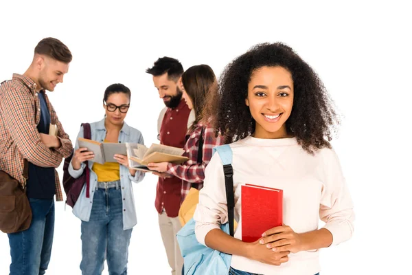 Atractiva chica afroamericana rizada sosteniendo libro cerca de grupo de jóvenes aislados en blanco - foto de stock