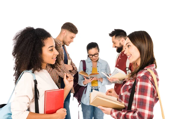 Meninas multiétnicas atraentes que falam com livros em mãos perto do grupo de jovens isolados no branco — Fotografia de Stock