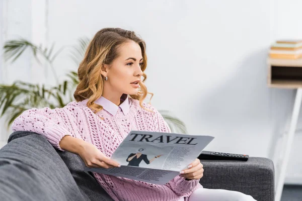 Hübsche Frau sitzt auf Sofa, hält Reisezeitung in der Hand und schaut im Wohnzimmer fern — Stockfoto