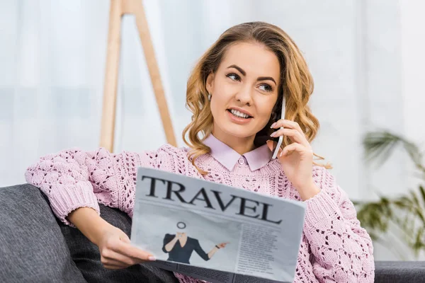 Mujer bonita sentada en el sofá, hablando en el teléfono inteligente y la celebración de periódico de viaje en la sala de estar - foto de stock