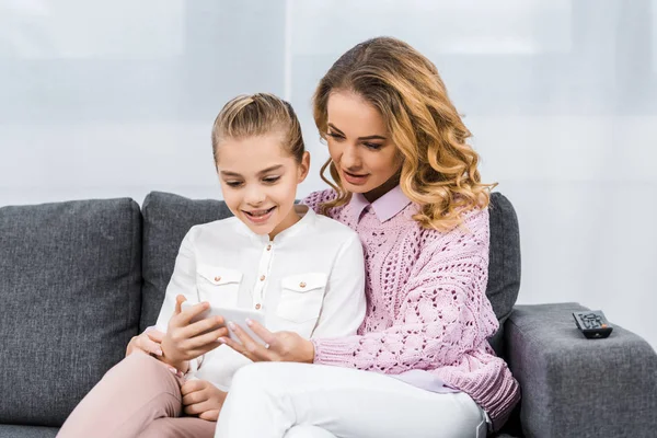 Hübsche Frau und nettes Mädchen sitzen auf Sofa und benutzen Smartphone im Wohnzimmer — Stockfoto