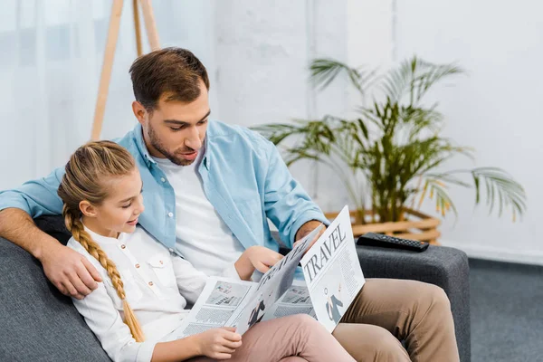 Красивий батько і мила дочка сидить на дивані і читає подорожі газети у вітальні — стокове фото