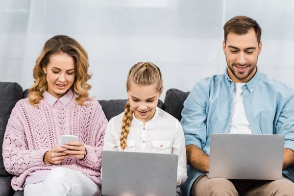 Mujer bonita sentada en el sofá y el uso de teléfono inteligente, mientras que la hija y el marido escribiendo en los teclados del ordenador portátil en la sala de estar - foto de stock