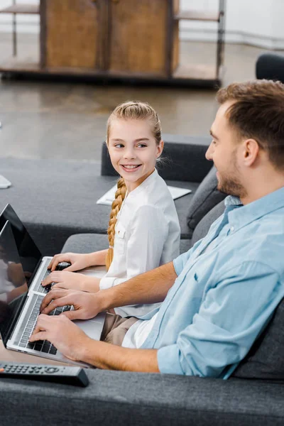 Smiling father and daughter sitting on sofa, typing on laptop keyboards and looking at each other — Stock Photo