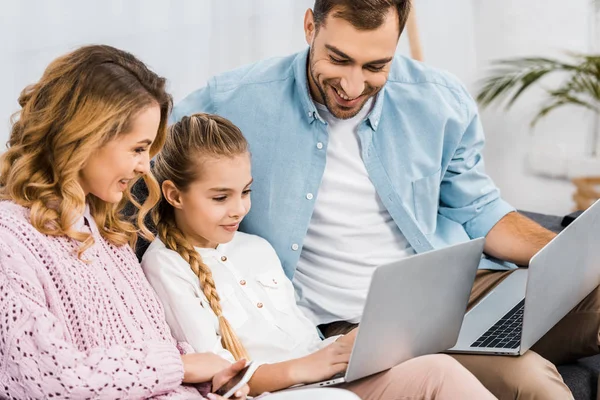 Smiling parents sitting on sofa and looking at laptop holding by cute girl — Stock Photo