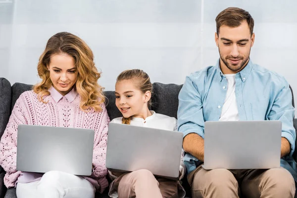 Cute girl with two parents sitting on sofa and using laptops in apartment — Stock Photo