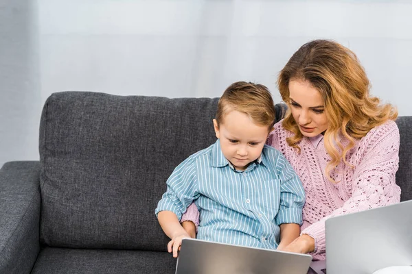 Bonita mujer con hijo sentado en el sofá y señalando con el dedo en la pantalla del ordenador portátil en el apartamento - foto de stock