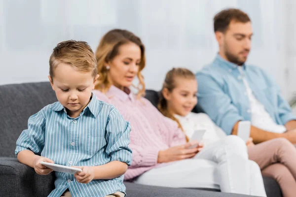 Mignon garçon assis sur le canapé et en utilisant smartphone avec la famille à l'arrière-plan dans le salon — Photo de stock