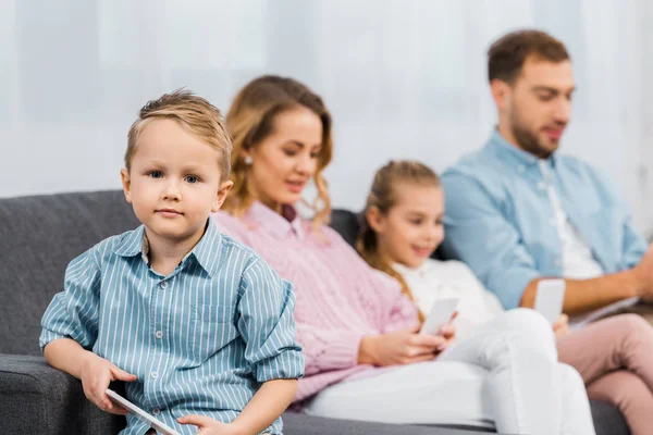 Симпатичный мальчик сидит на диване, держит смартфон и смотрит на камеру с семьей на заднем плане в квартире — стоковое фото