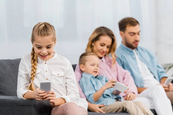 Menina bonito sentado no sofá e usando smartphone com a família no fundo na sala de estar — Fotografia de Stock