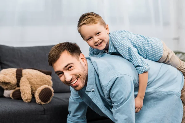 Fröhlicher Junge, der auf dem Rücken seines Vaters sitzt, auf allen Vieren steht und in Wohnung in die Kamera schaut — Stockfoto