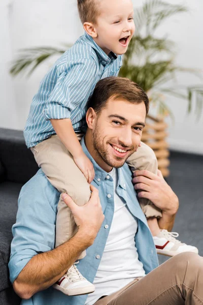 Niño sonriente sentado en los hombros del padre mirando la cámara en la sala de estar - foto de stock