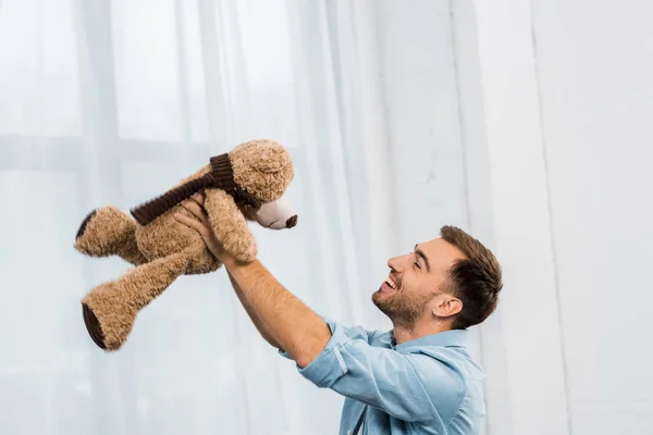 Sonriente hombre sosteniendo oso de peluche en levantar las manos en la sala de estar - foto de stock