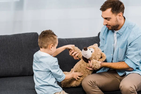 Sonriente padre e hijo sentado en el sofá y sosteniendo oso de peluche en la sala de estar - foto de stock