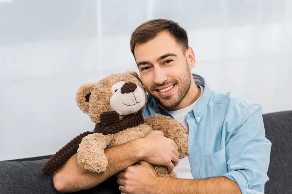 Homem bonito sorrindo, abraçando ursinho de pelúcia e olhando para a câmera no apartamento — Fotografia de Stock