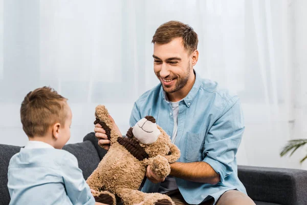 Smiling father and son sitting on sofa and holding teddy bear in living room — Stock Photo