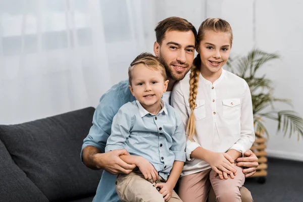 Улыбающийся отец с милыми детьми, сидящими на диване и смотрящими на камеру в гостиной — стоковое фото