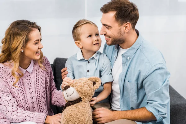 Cheerful parents sitting on sofa and embracing cute son holding teddy bear in apartment — Stock Photo