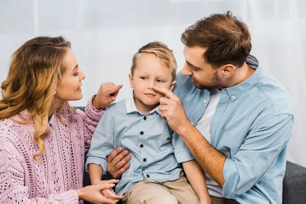 Happy parents sitting on sofa and touching with fingers nose of son in apartment — Stock Photo