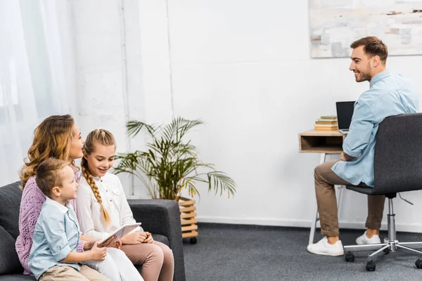 Hübsche Frau mit Kindern auf Sofa sitzend und Ehemann am Tisch in Wohnung anschauend — Stockfoto