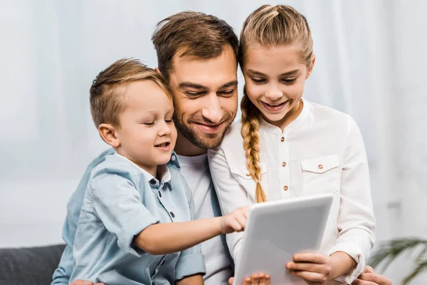 Padre sonriente abrazando linda chica sosteniendo tableta digital y adorable chico apuntando con el dedo a la pantalla en la sala de estar - foto de stock