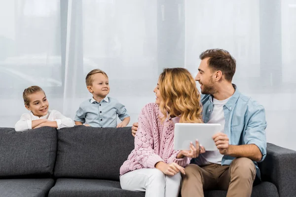 Eltern sitzen auf Sofa, halten digitales Tablet in der Hand und schauen Kinder im Wohnzimmer an — Stockfoto