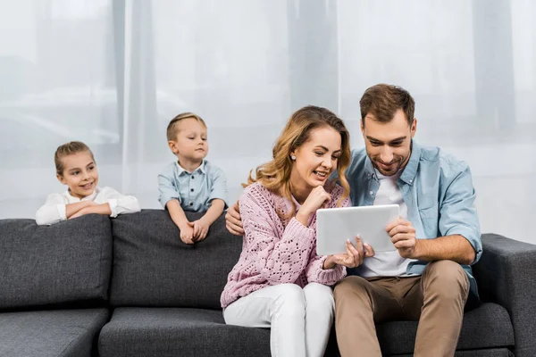 Nette Geschwister betrachten Eltern, die auf dem Sofa sitzen und digitales Tablet im Wohnzimmer benutzen — Stockfoto