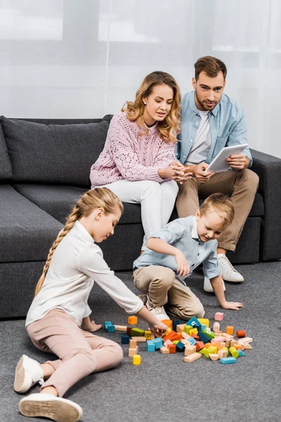 Parents assis sur le canapé avec tablette numérique et regardant les enfants jouer avec des blocs de bois multicolores sur le sol dans l'appartement — Photo de stock