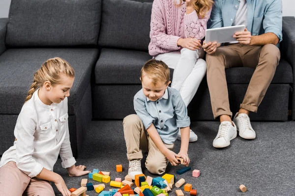 Vue recadrée des parents assis sur le canapé et utilisant une tablette numérique pendant que les enfants jouent avec des blocs de bois multicolores sur le sol dans le salon — Photo de stock