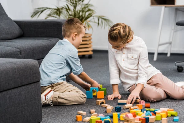 Bonito menina e menino sentado no chão e brincando com blocos de madeira multicoloridos no apartamento — Fotografia de Stock