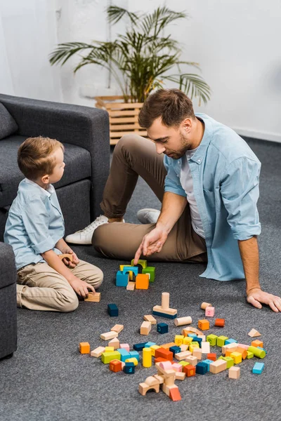 Handsome father and cute son playing with multicolored wooden blocks on floor in living room — Stock Photo