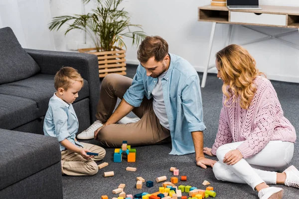 Mãe, pai e filho bonito brincando com blocos de madeira multicoloridos no chão na sala de estar — Fotografia de Stock