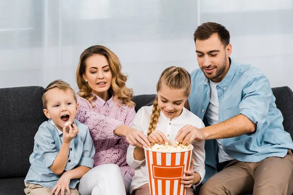 Happy family sitting on sofa, watching tv and eating popcorn from striped bucket in living room — Stock Photo