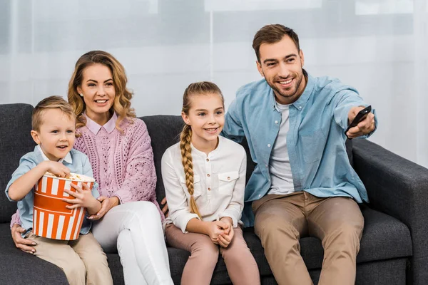 Bonito pai sentado no sofá e mudando de canais por controle remoto com sorridente esposa e filha e filho comendo pipocas no apartamento — Fotografia de Stock