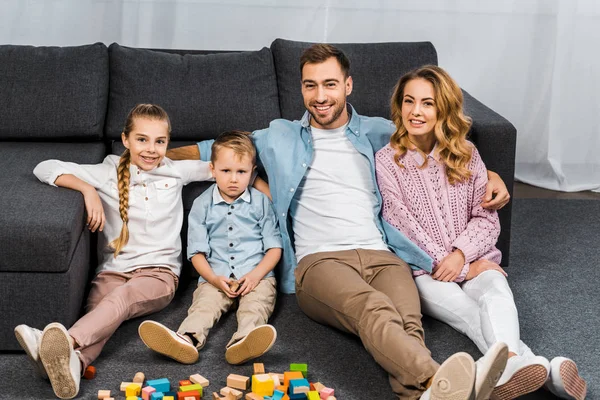Padre sonriente, madre, hija e hijo molesto sentado en el piso cerca del sofá en el apartamento - foto de stock