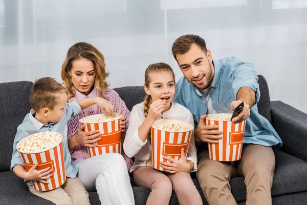 Smiling father sitting on sofa and changing channels by remote controller with family holding striped buckets and eating popcorn in apartment — Stock Photo