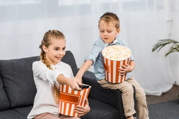 Lächelndes Mädchen, das auf dem Sofa sitzt und Kanäle mit Fernbedienung wechselt, während Bruder Popcorn in der Wohnung isst — Stockfoto