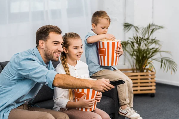 Padre sorridente seduto sul divano e cambiando canale con telecomando con bambini che tengono secchi di popcorn a strisce in appartamento — Foto stock