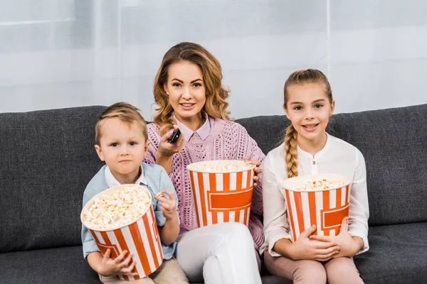 Lächelnde Mutter mit Kindern, die auf dem Sofa sitzen, gestreifte Popcorn-Eimer in der Hand halten und per Fernbedienung im Wohnzimmer die Kanäle wechseln — Stockfoto
