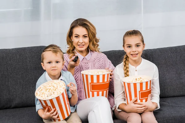 Mère souriante avec des enfants assis sur le canapé, tenant des seaux de maïs soufflé rayé et changer de canaux par télécommande dans le salon — Photo de stock