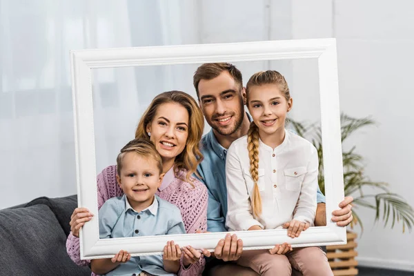 Família feliz segurando moldura branca e olhando para a câmera na sala de estar — Fotografia de Stock