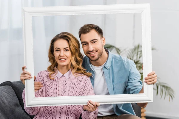 Smiling man and woman holding white frame and looking at camera in living room — Stock Photo